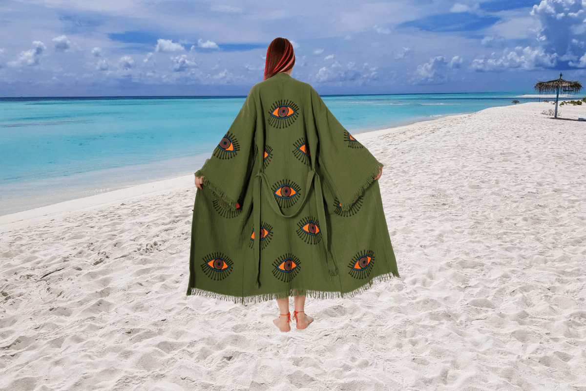 a woman standing barefoot on a beach at sunset, wearing a flowing beige kimono robe with blue evil eye prints. The robe billow