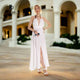 A stunning woman radiates elegance in a boho white cotton maxi dress, standing gracefully in front of a luxurious hotel.