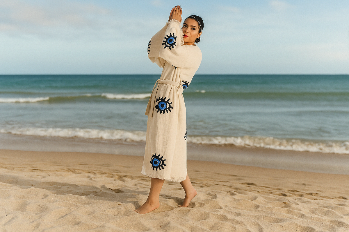 A woman wearing a flowing evil‑eye printed kimono cover‑up standing confidently on a sunny beach, the sea breeze ruffling the 