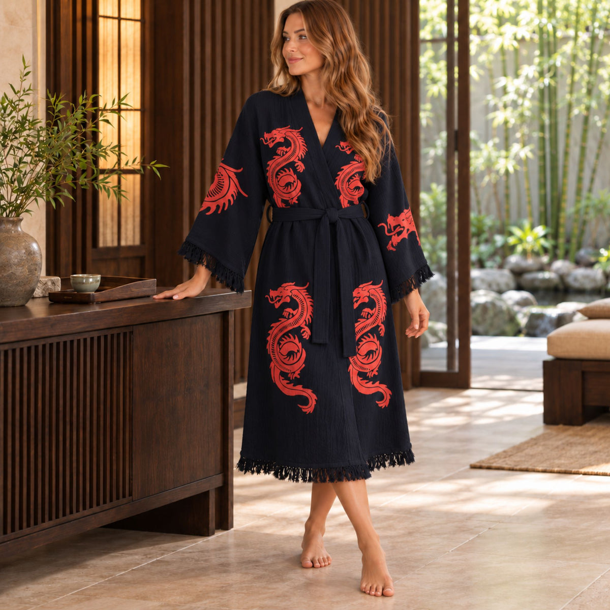 Woman standing beside a wooden console in a black kimono robe with red dragon prints, a tied waist belt and fringed cuffs and hem.